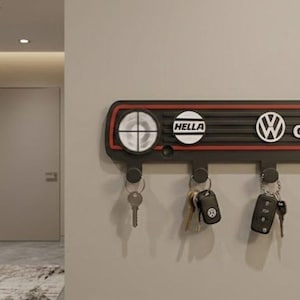 Puede incluir: Llavero negro y rojo con el texto "GTI" y "HELLA". El llavero tiene cinco ganchos con llaves colgando. El llavero está montado en una pared de color beige claro.
