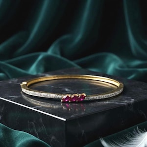 Puede incluir: Una pulsera rígida articulada de tono dorado con una hilera de pequeñas piedras transparentes tipo pavé a lo largo de la banda y tres gemas de color rojo rosado intenso en talla marquesa dispuestas horizontalmente en la parte frontal.