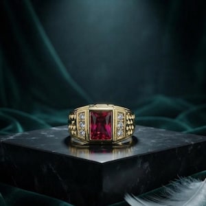 May include: A gold ring featuring a central rectangular red gemstone flanked by two vertical rows of three small clear stones on each side. The band has a textured, geometric pattern on the sides, providing a detailed and structured appearance.