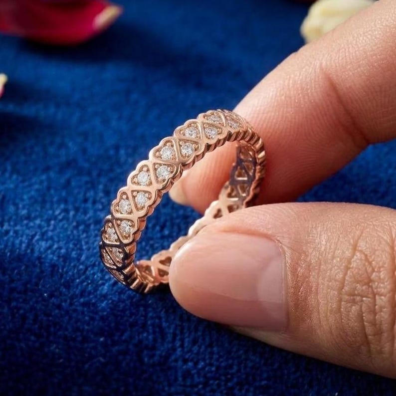 May include: Rose gold ring with a heart-shaped design, set with small, sparkling diamonds. The ring is held against a blue background, showcasing its intricate details and elegant style.