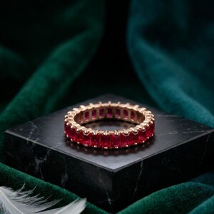 May include: An eternity band ring featuring a continuous row of vibrant red, rectangular-cut stones set in a polished gold-tone metal band. The ring is positioned on a smooth, black marble block, showcasing the uniform alignment of the gemstones.