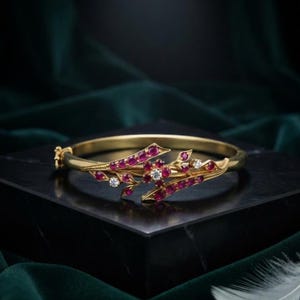 Puede incluir: Un brazalete rígido de oro con bisagra que presenta un diseño de inspiración floral adornado con múltiples rubíes pequeños de color rojo intenso y varios diamantes transparentes de talla redonda engastados a lo largo de las ramas de metal.
