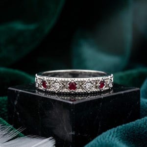 May include: A silver-toned band ring featuring an alternating pattern of square-cut red rubies and round-cut clear stones set within a textured, beaded metal frame. The ring displays a repeating geometric design across its entire circumference.
