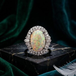 May include: A silver-toned ring featuring a large, oval-shaped iridescent opal center stone. The opal displays flashes of green, orange, and yellow. It is surrounded by a double halo of small, clear, round and baguette-cut white diamonds.