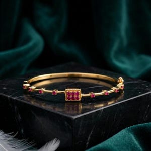 Puede incluir: Una pulsera rígida de tono dorado con un engaste central rectangular incrustado con seis pequeñas piedras rojas vibrantes. La banda está acentuada con piedras rojas individuales adicionales espaciadas uniformemente a lo largo del marco metálico.