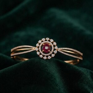 Op de afbeelding: Een gouden armband met een centraal cirkelvormig ontwerp. Het midden bevat een ronde rode edelsteen omringd door twee concentrische ringen van kleine witte pavé-gezette stenen. De band splitst zich in twee dunne, met steentjes bezette armen.