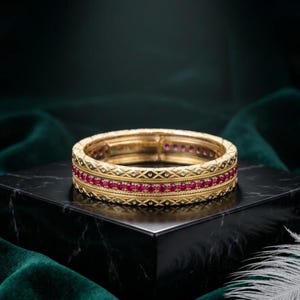May include: A gold band ring featuring a central row of small, round, vibrant red rubies. The metal surface is decorated with a repeating diamond-shaped lattice pattern and fine beaded detailing along the edges, creating a textured appearance.