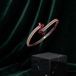 May include: A rose gold hinged bangle bracelet featuring two parallel rows of small white pavé diamonds and a central floral cluster composed of four pear-shaped deep red rubies set in a bypass design.