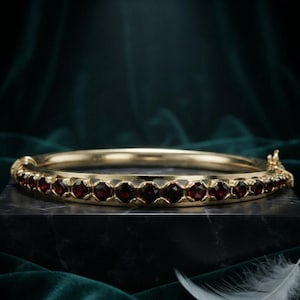 May include: A gold hinged bangle bracelet featuring a continuous row of round, deep red garnet gemstones set in a metallic frame. The bracelet has a secure clasp mechanism on one side and a smooth, polished gold finish along the inner band.