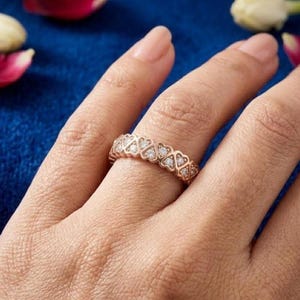 May include: A rose gold ring featuring a repeating heart design, each heart set with small, clear gemstones. The ring is worn on a finger, with a blue background and flower petals visible.