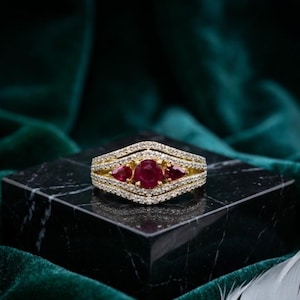 Könnte beinhalten: Ein Goldring mit einem zentralen, rund geschliffenen roten Rubin, flankiert von zwei tropfenförmig geschliffenen roten Rubinen. Die Ringschiene ist zweigeteilt und mit kleinen weißen Pavé-Diamanten besetzt, die der V-Form der Fassung folgen.