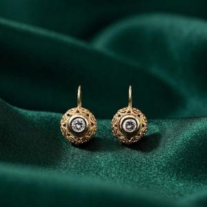 May include: A pair of gold drop earrings featuring a spherical design with intricate filigree metalwork and a single round brilliant-cut white gemstone set in the center of each sphere, suspended from a simple gold hook mechanism.