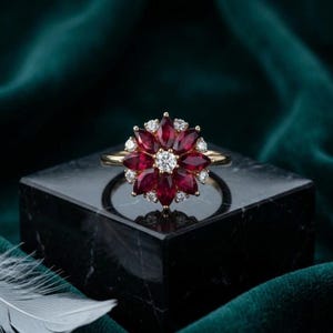 Puede incluir: Un anillo de oro con un diamante blanco redondo central rodeado por un arreglo floral de rubíes de color rojo intenso en talla marquesa y diamantes blancos redondos más pequeños, creando un diseño de racimo que recuerda a una flor.
