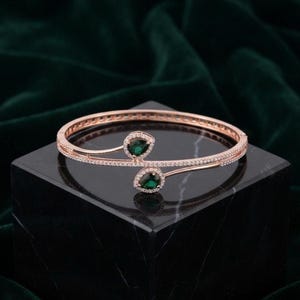 May include: A rose gold hinged bangle bracelet featuring two teardrop-shaped green stones at the open ends, surrounded by small clear pavé stones along the entire band and the settings of the green stones.