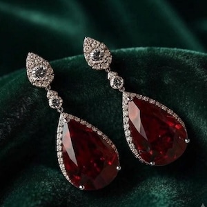 May include: A pair of drop earrings featuring large, pear-shaped, deep red faceted stones. Each earring is framed by a border of small, clear, sparkling stones and includes two additional small, round, clear stones set along the vertical metal post.