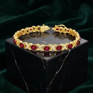 May include: A gold-tone bracelet featuring a repeating X-shaped link pattern alternating with oval-cut red stones. The metal links are accented with small clear stones. The clasp is engraved with the text IEFIL. The bracelet is circular in shape.