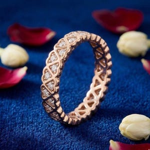 May include: Rose gold eternity ring with a repeating pattern of diamond-studded shapes. The ring is set against a deep blue background, with scattered rose petals and white flower buds.