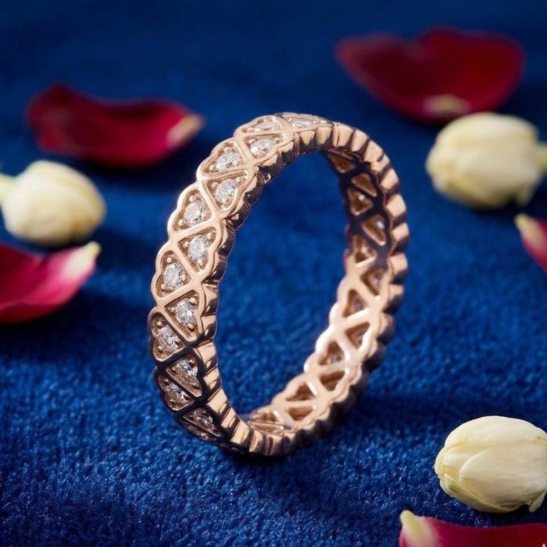 May include: Rose gold eternity ring with a repeating pattern of diamond-studded shapes. The ring is set against a deep blue background, with scattered rose petals and white flower buds.