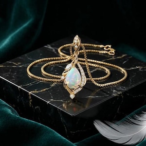 May include: A gold necklace featuring a teardrop-shaped opal pendant surrounded by small, clear, round-cut stones. The pendant is set in a gold frame with intricate, swirling metalwork, and the chain is a delicate gold box link style.