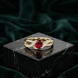 Op de afbeelding: Een gouden ring met een centrale ovaal geslepen rode edelsteen, geflankeerd door twee in elkaar grijpende banden. Elke band is bezet met een rij kleine, heldere, rond geslepen stenen die vanaf de zetting langs de gouden ring doorlopen.