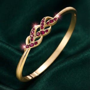 Pode incluir: Uma pulseira rígida de ouro com um elemento central de design trançado. A seção trançada é adornada com várias pequenas pedras preciosas redondas de cor vermelho profundo incrustadas no metal, criando um padrão contínuo e complexo.