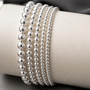 May include: A set of five silver-toned beaded bracelets arranged in a row. Each bracelet features a different size of polished, spherical beads, ranging from larger beads on the left to smaller, finer beads on the right, creating a graduated look.