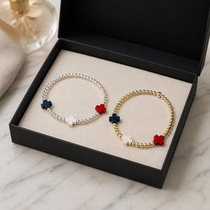 Puede incluir: Dos pulseras de cuentas presentadas en una caja de regalo negra. Una pulsera tiene cuentas plateadas con tres dijes en forma de cruz en azul marino, blanco y rojo. La segunda pulsera consta de cuentas doradas con cruces a juego en azul, blanco y rojo.