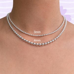 May include: Two silver ball chain necklaces, one with 3mm beads and the other with 4mm beads, are displayed around a person's neck. The text "3mm" and "4mm" are visible, indicating the bead sizes. The necklaces are a simple, elegant jewelry.