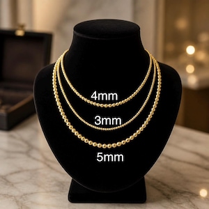 May include: Three gold-toned beaded necklaces displayed on a black bust. The necklaces feature spherical beads in three distinct sizes, labelled with text indicating diameters of 4mm, 3mm, and 5mm, showing the variation in bead size for each chain.