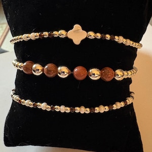 May include: Three beaded bracelets featuring metallic gold-tone spheres, small dark brown translucent beads, and shimmering copper-colored stone beads. The top bracelet includes a central four-petaled flower-shaped charm in a polished gold-tone finish.