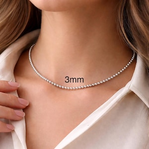 May include: A delicate silver necklace with 3mm beads. The necklace is worn around the neck, resting on a white shirt. The text "3mm" is displayed on the image.