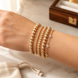 May include: A stack of five gold-toned bracelets worn on a wrist. The set includes three beaded bracelets with varying bead sizes, one thin chain bracelet with small beads, and one delicate chain bracelet featuring four-petaled flower charms.