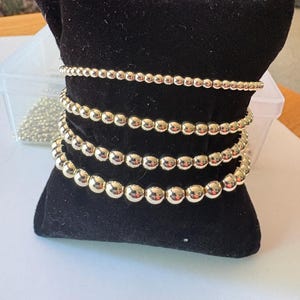 May include: Four stretch bracelets made of polished gold-tone spherical beads of varying sizes, arranged in a stack on a black velvet display pillow. The beads increase in diameter from the top bracelet to the bottom bracelet.