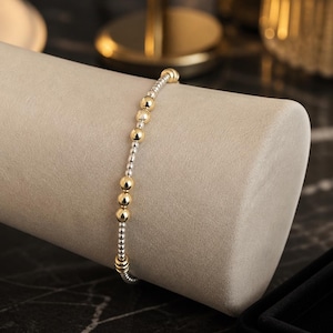 May include: A beaded bracelet featuring a repeating pattern of small silver-toned metallic spheres interspersed with larger, polished gold-toned metallic spheres. The beads are strung on a flexible cord, creating a continuous circular design.