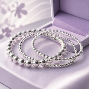 Puede incluir: Tres pulseras de cuentas plateadas presentadas en una caja de joyería morada. Las pulseras varían en tamaño de cuentas, con las cuentas más grandes en el centro de la pulsera delantera. La caja está abierta, revelando un interior de satén.