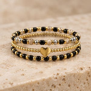 May include: A set of three stacked bracelets featuring small round beads. The top bracelet has alternating black, silver, and beige beads. The middle bracelet consists of gold beads with a central gold heart charm. The bottom bracelet has black and gold beads.