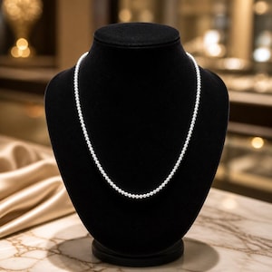 May include: A delicate pearl necklace displayed on a black velvet bust. The necklace features numerous small, round, white pearls. The bust is set against a blurred background with warm tones, highlighting the jewelry's elegance.