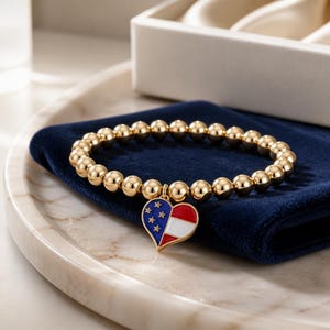 May include: A gold-toned beaded bracelet featuring a heart-shaped charm. The charm is enamelled with a blue section containing white stars and red and white stripes, mimicking the American flag design. The beads are round, polished, and metallic.