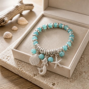 May include: A stretch bracelet featuring round, light blue beads interspersed with small, faceted silver-toned metal spacers. Three silver-toned charms hang from the bracelet: a textured seashell, a mermaid figure, and a five-pointed starfish.