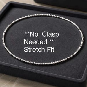 May include: A silver beaded necklace with a stretch fit design. The necklace is displayed on a dark gray surface. Text on the image reads "No Clasp Needed" and "Stretch Fit".