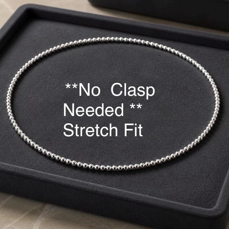 May include: A silver beaded necklace with a stretch fit design. The necklace is displayed on a dark gray surface. Text on the image reads "No Clasp Needed" and "Stretch Fit".