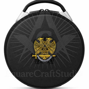 Puede incluir: Estuche redondo negro con asa y detalles plateados. El estuche presenta un emblema dorado con dos águilas, el número 32 y el texto "SPES MEA IN DEO EST". Un símbolo masónico es sutilmente visible en el fondo.