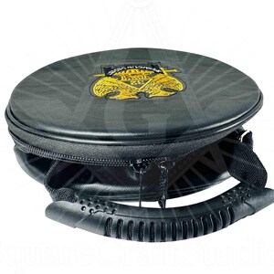 May include: A black, round carrying case with a zipper closure and a handle. The top of the case features a gold emblem with the text "PERMEA IN OBRAS". The case is designed to carry a round object.