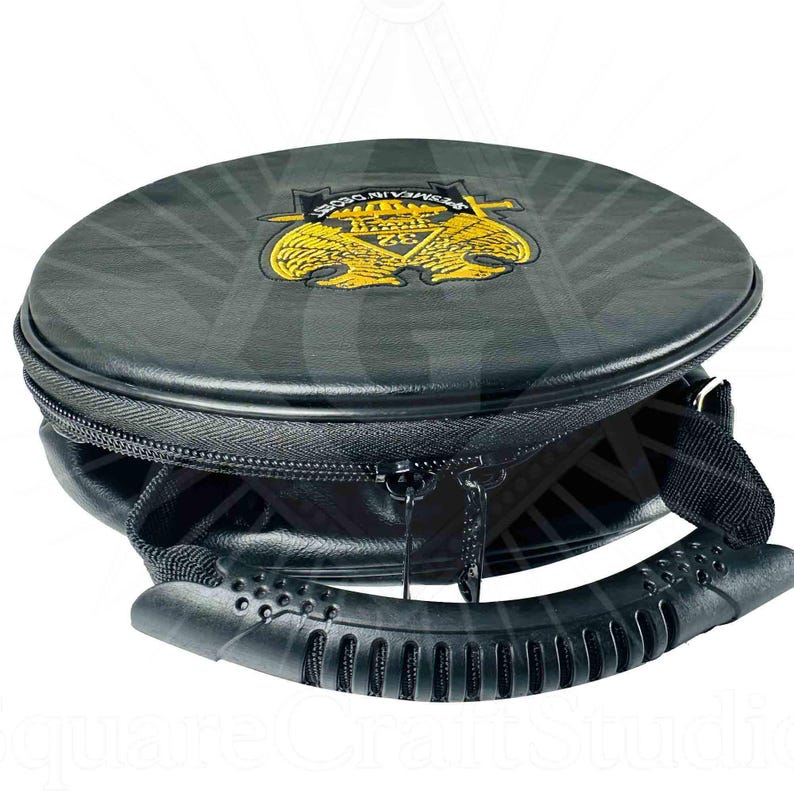 May include: A black, round carrying case with a zipper closure and a handle. The top of the case features a gold emblem with the text "PERMEA IN OBRAS". The case is designed to carry a round object.