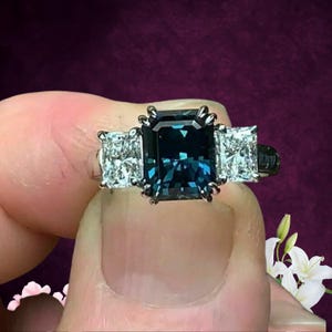 May include: A three-stone ring featuring a large, emerald-cut, dark teal gemstone flanked by two smaller, square, clear gemstones. The ring has a silver band and is set against a dark purple background.