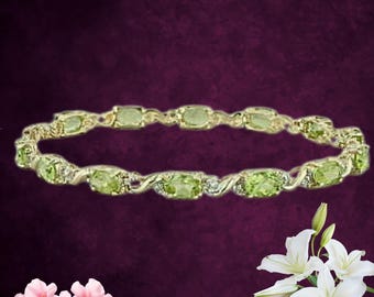 May include: A gold-toned bracelet featuring oval-shaped, light green gemstones. The bracelet has a delicate, interwoven design with small, clear accent stones. The background is a deep purple, with pink and white flowers in the corners.