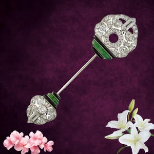 Könnte beinhalten: Eine silberfarbene Anstecknadel mit zwei dekorativen Enden, die mit klaren runden und baguettegeschliffenen Steinen besetzt sind und durch Bänder aus schwarzer und grüner Emaille akzentuiert werden. Ein langer, dünner Metallschaft verbindet die Köpfe.