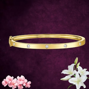 Op de afbeelding: Een gouden armband met zes ronde, heldere edelstenen gelijkmatig verdeeld. De armband heeft een sluiting en staat tegen een donkerpaarse achtergrond. Roze en witte bloemen zijn op de voorgrond.
