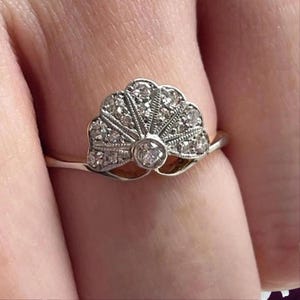 May include: A close-up of a ring with a fan-shaped design. The ring features a central, round diamond and numerous smaller diamonds set in a silver-colored metal. The fan design is detailed and intricate.