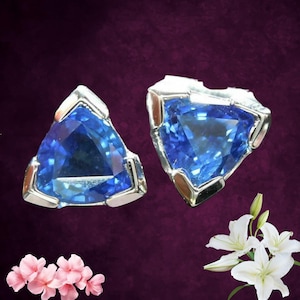 Könnte beinhalten: Zwei dreieckige blaue Edelsteinohrringe mit silber- und goldfarbenen Metalleinfassungen. Die Edelsteine sind leuchtend blau. Die Ohrringe werden vor einem dunkelvioletten Hintergrund mit rosa und weißen Blumen im Vordergrund präsentiert.